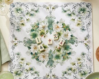 May include: A square mahjong mat featuring a white background with a central circular arrangement of white and green floral illustrations, surrounded by ornate black line-art borders. Four ivory mahjong tiles with red and black characters are placed in the center.