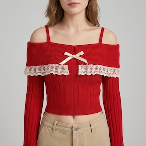 Puede incluir: Una blusa de punto acanalado de color rojo brillante con mangas largas y diseño de hombros descubiertos con tirantes finos. La prenda está adornada con un borde de encaje color crema en el escote y un pequeño lazo de cinta color crema en el centro.