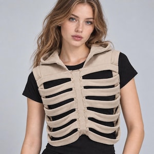 Puede incluir: Un chaleco beige sin mangas con capucha que presenta un diseño de caja torácica con tiras de tela horizontales en la parte delantera, usado sobre una camiseta negra lisa de manga corta. El chaleco tiene una apariencia estructurada.