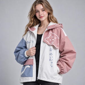 Könnte beinhalten: Eine farblich abgesetzte Reißverschlussjacke mit weißer Mitte, einem altrosa linken Ärmel und Kapuzenbereich sowie einem gedeckten blauen rechten Ärmel und einer Tasche. Die Jacke zeigt weiße Linienzeichnungen eines Katzengesichts, eines Schwanzes und japanischer Schriftzeichen.