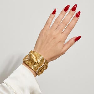 Puede incluir: Un brazalete ancho de tono dorado con una superficie texturizada y estriada, motivos de conchas en relieve y delicados detalles de cuentas blancas. El brazalete tiene un acabado metálico y un diseño estructurado que rodea la muñeca.
