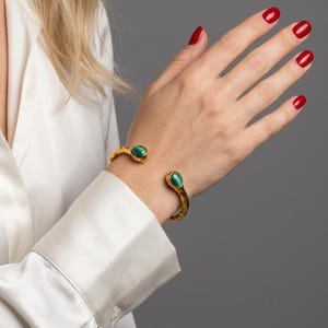 Può includere: Un bracciale a polsino color oro caratterizzato da una superficie metallica testurizzata e organica, con due pietre cabochon ovali verdi incastonate alle estremità aperte. Il bracciale presenta una finitura lucida e un design a fascia larga.