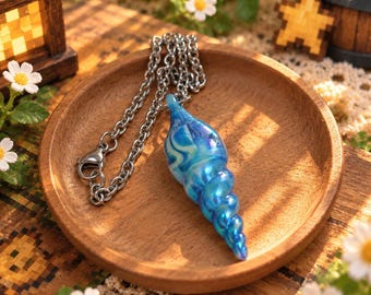 May include: A spiral-shaped pendant made of iridescent blue and white glass, attached to a silver-toned metal chain. The pendant features a swirling, translucent pattern that shifts in color, resting on a circular wooden tray.