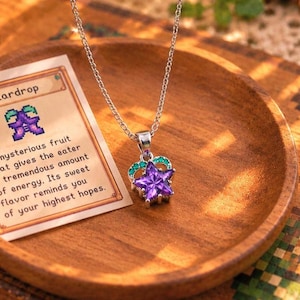 May include: A silver-toned necklace featuring a star-shaped purple pendant with small green accents. Beside it lies a rectangular card with the text Stardrop and a description of a mysterious fruit that gives the eater energy.