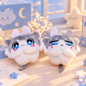 May include: Two plush toys featuring grey and white fur, large blue eyes, and small ears. One plush has wide, bright blue eyes, while the other has narrowed, patterned blue eyes. Both have pink cheek accents and small white paws.