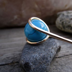 May include: A turquoise gemstone wrapped in silver wire, set on a grey stone. The gemstone is round and has a smooth surface. The wire is twisted around the gemstone and forms a loop at the top.