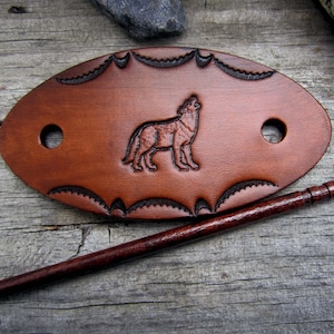 Puede incluir: Un palo para el pelo de cuero marrón con un diseño de lobo y un pasador de madera. El cuero está decorado con un borde festoneado.