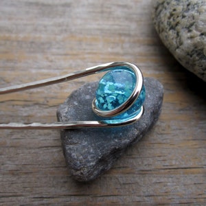 May include: A silver hair pin with a hammered texture, featuring a light blue glass orb with a speckled design. The orb is encircled by a silver wire. The pin is resting on a gray stone, set against a wooden background.