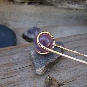 May include: A gold wire wrapped around a round, purple amethyst stone, attached to a long, thin gold hair stick.
