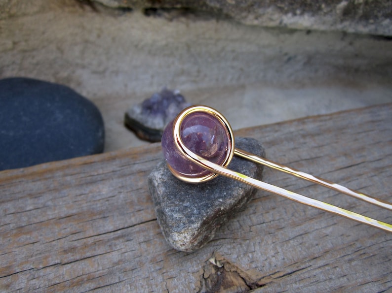 May include: A gold wire wrapped around a round, purple amethyst stone, attached to a long, thin gold hair stick.