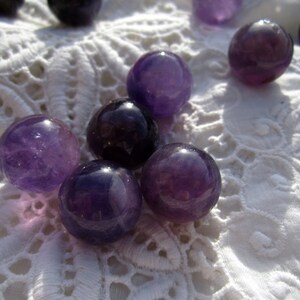 May include: A group of seven amethyst gemstone spheres, ranging in size, are displayed on a white lace surface. The spheres are a deep purple color and have a smooth, polished finish.