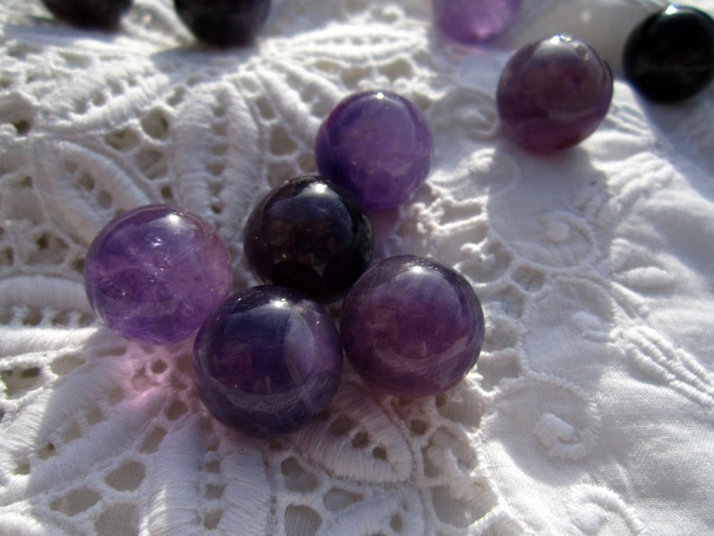 May include: A group of seven amethyst gemstone spheres, ranging in size, are displayed on a white lace surface. The spheres are a deep purple color and have a smooth, polished finish.