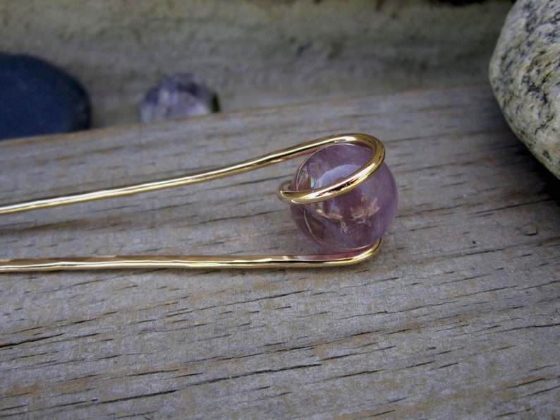 May include: A gold hair stick with a purple amethyst gemstone sphere at the end. The sphere is encased in a gold wire cage.