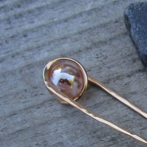 Puede incluir: Un palillo para el pelo de color dorado con una esfera rosa nacarada en el extremo. El palillo est&aacute; martillado y tiene un acabado texturizado.