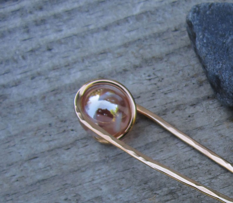 Puede incluir: Un palillo para el pelo de color dorado con una esfera rosa nacarada en el extremo. El palillo est&aacute; martillado y tiene un acabado texturizado.