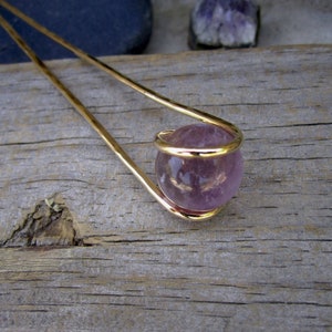 May include: A gold hair stick with a purple amethyst sphere at the end. The sphere is encased in a gold wire cage.