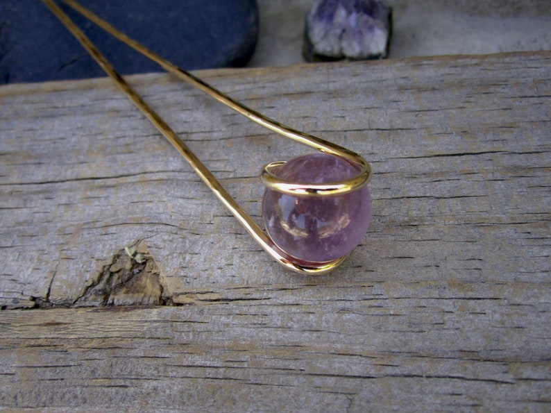 May include: A gold hair stick with a purple amethyst sphere at the end. The sphere is encased in a gold wire cage.