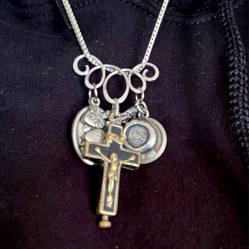 May include: A silver chain necklace with a silver and black crucifix pendant, and several other silver charms. The charms include a heart, a small cross, and a round charm with an image of a bird.