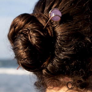 May include: A person with brown hair in a bun, wearing a purple shirt, with a hair clip with a purple stone.