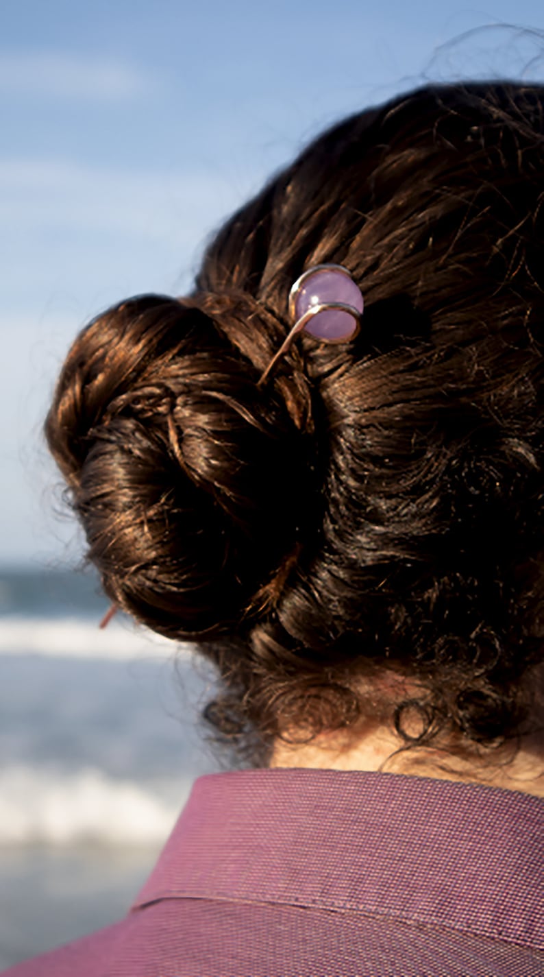 May include: A person with brown hair in a bun, wearing a purple shirt, with a hair clip with a purple stone.