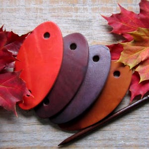 May include: Five leather swatches in various shades of red, brown, purple, and orange. The swatches are oval-shaped with two holes punched in each. A wooden dowel is lying on top of the swatches.