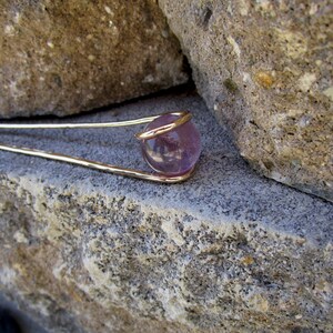 May include: A gold wire wrapped around a round, purple amethyst gemstone. The gemstone is set against a backdrop of rough, gray stone.