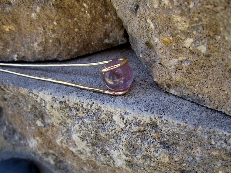 May include: A gold wire wrapped around a round, purple amethyst gemstone. The gemstone is set against a backdrop of rough, gray stone.