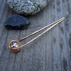 Puede incluir: Una varita para el pelo de oro rosa con una cuenta de vidrio transparente en el extremo. La varita est&aacute; sobre una superficie de madera.