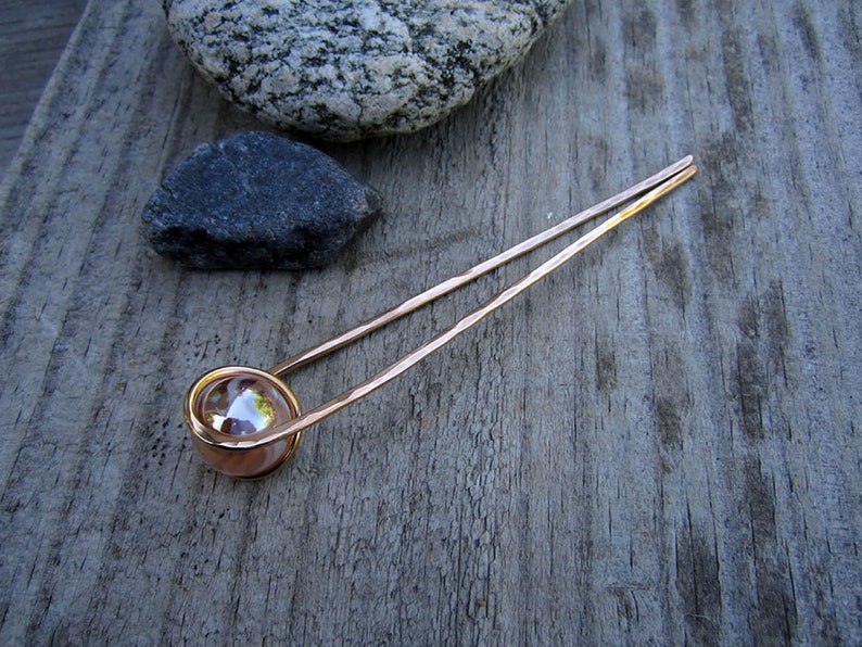 Puede incluir: Una varita para el pelo de oro rosa con una cuenta de vidrio transparente en el extremo. La varita est&aacute; sobre una superficie de madera.