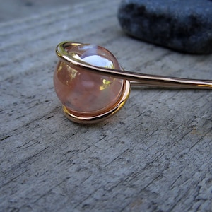 Puede incluir: Una varita para el pelo dorada con una esfera de vidrio rosa iridiscente en el extremo. La esfera est&aacute; encerrada en un bucle de alambre.