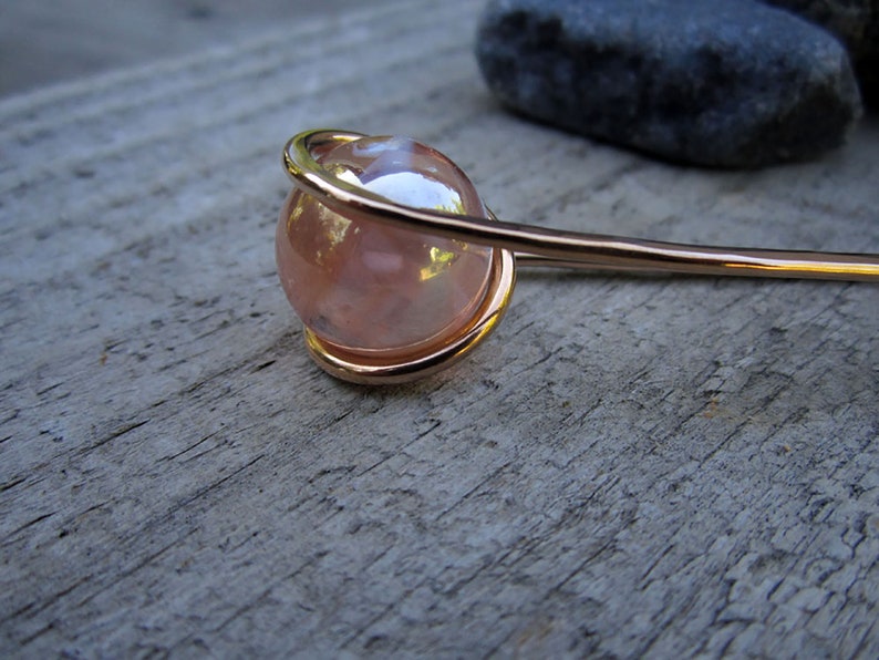 Puede incluir: Una varita para el pelo dorada con una esfera de vidrio rosa iridiscente en el extremo. La esfera est&aacute; encerrada en un bucle de alambre.