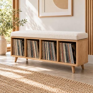 May include: A rectangular wooden storage bench with a light beige upholstered seat cushion. The base features four tapered wooden legs and four open square compartments, each filled with a collection of vinyl records standing upright in rows.