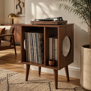 May include: A dark wood record player stand with four tapered legs, featuring two open storage compartments. The left side holds a collection of vinyl records, while the right side contains books and a small wooden bowl. A turntable sits on top.