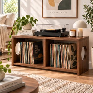 May include: A rectangular wooden media console with two open storage compartments filled with vinyl records. The unit features circular cutouts on the side panels and a smooth top surface supporting a black turntable, a candle, and a lamp.