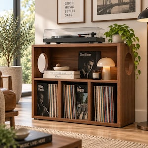 Puede incluir: Un mueble de madera para discos con acabado en nogal oscuro, que presenta dos aberturas laterales circulares, una superficie superior para un tocadiscos y tres compartimentos inferiores llenos de discos de vinilo, libros y una lámpara de hongo.