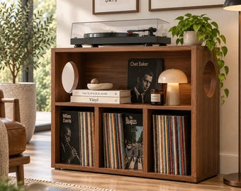 May include: A rectangular wooden record storage cabinet with a dark walnut finish, featuring two circular side cutouts, a top surface for a turntable, and three lower compartments filled with vinyl records, books, and a mushroom-shaped lamp.