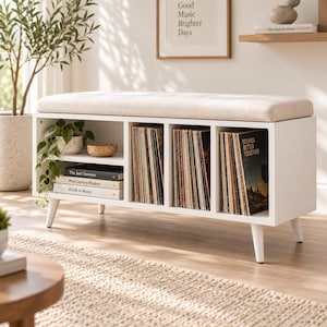 May include: A white storage bench with a beige upholstered seat cushion, featuring three vertical compartments filled with vinyl records and a side section with two shelves holding a small plant, a bowl, and books titled The Jazz Sessions, Mid-Century Modern, and A Life in Music.