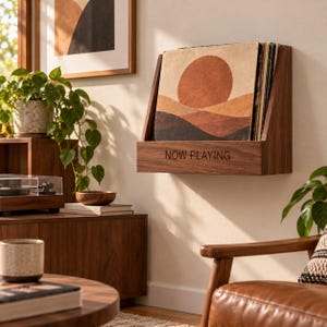 Peut inclure: Une étagère murale en bois foncé pour disques vinyles, dotée d'un dossier incliné et d'un panneau avant gravé avec le texte NOW PLAYING. L'étagère contient une pochette de disque vinyle avec un imprimé graphique d'un soleil sur des collines.