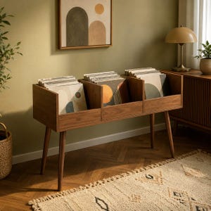 Op de afbeelding: Een houten opbergmeubel voor vinylplaten met drie open vakken, ondersteund door vier taps toelopende poten. De kast is gevuld met diverse vinylplaten met abstracte geometrische hoezen in de kleuren oranje, blauw en crème.