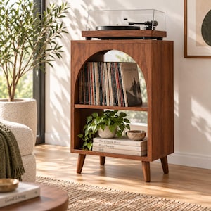 May include: A dark wood record player stand featuring an arched open shelf design with four tapered legs. The top surface holds a black turntable with a clear dust cover. The middle shelf contains a row of vinyl records and a green potted plant.