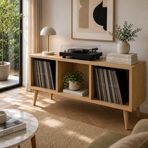 May include: A light oak wood media console with three open storage compartments, supported by four tapered legs. The unit holds two rows of vinyl records, a black turntable, a white table lamp, a potted plant, and stacked books.