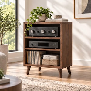 May include: A dark wood media console with three open shelves supported by four tapered legs. The top shelf holds a black audio amplifier, the middle shelf a black media player, and the bottom shelf a collection of vinyl records and a woven basket.