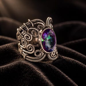 May include: A silver-toned wire-wrapped ring featuring an oval-shaped, multi-colored iridescent gemstone. The metal band is constructed from intricate, swirling wire patterns that create an openwork design surrounding the central stone.