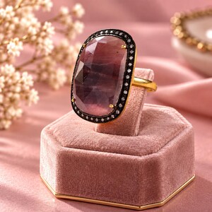 May include: A gold ring featuring a large, oval-shaped, faceted pink gemstone. The stone is surrounded by a dark, oxidised metal border encrusted with small, clear white stones. The ring is displayed on a pink velvet stand.