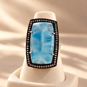 May include: A rectangular ring featuring a large, smooth, light blue stone with natural white and grey marbling. The stone is set in a dark metal frame bordered by a row of small, clear, sparkling stones held in place by four silver prongs.