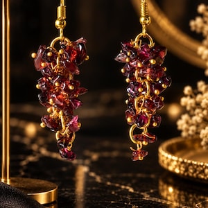 May include: A pair of dangle earrings featuring clusters of deep red, irregular garnet gemstone chips attached to gold-toned metal wire loops. The earrings hang from gold-toned fishhook ear wires, creating a textured, grape-like appearance.