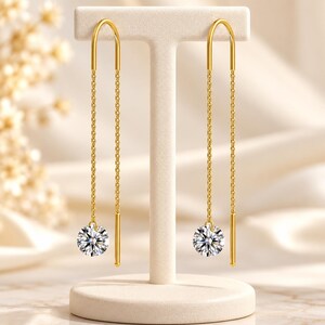 May include: A pair of gold threader earrings featuring a long, slender chain design. Each earring has a polished gold bar at the top and a single, round, brilliant-cut clear gemstone dangling at the end of the delicate gold chain.