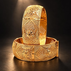May include: Two gold bangles featuring intricate filigree and openwork patterns. Each bracelet displays a repeating circular floral motif surrounded by textured geometric detailing and a polished finish, with a hinged clasp mechanism visible.