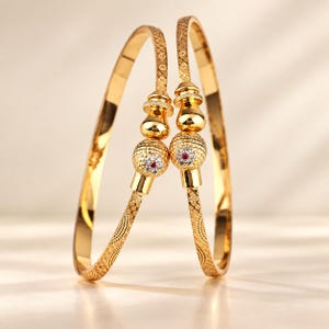 Puede incluir: Dos brazaletes dorados con un patrón de rayado cruzado texturizado en la banda. Cada brazalete está adornado con un elemento decorativo central que consiste en una cuenta dorada lisa y una esfera texturizada con una piedra roja y cristales.