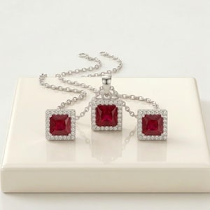 May include: A jewellery set consisting of a silver chain necklace with a square pendant and matching square stud earrings. Each piece features a deep red square-cut gemstone surrounded by a border of small white stones set in silver metal.
